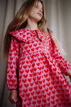 Roze Adore jurk met rode hartjesprint van Daily Brat, met rucheskraag en lange mouwen – gemaakt van biologisch katoen.